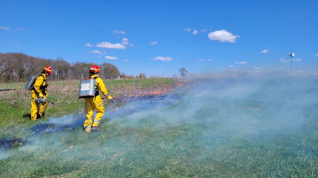 Two people in yellow uniforms lighting a fire on a prairie.